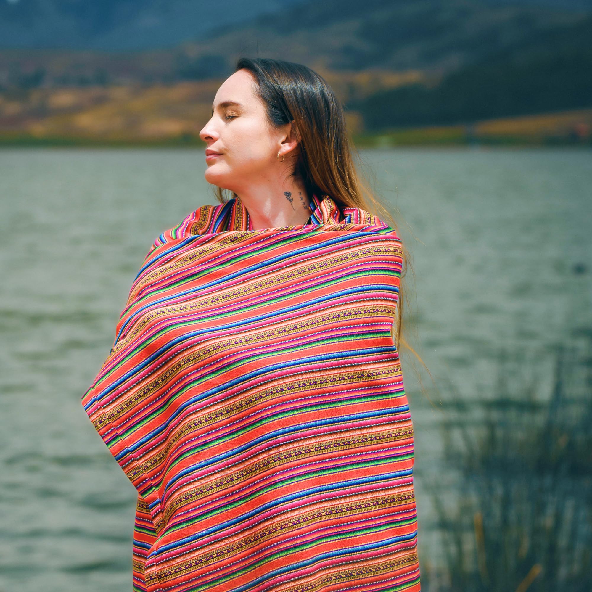 Woman wrapped in a colorful Peruvian woven blanket standing by a lake in a natural landscape