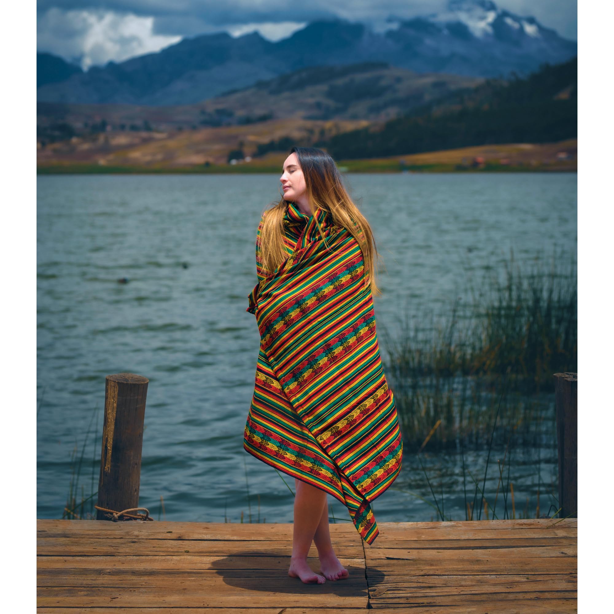 Woman wrapped in a colorful blanket standing on a wooden dock by a lake with mountains in the background