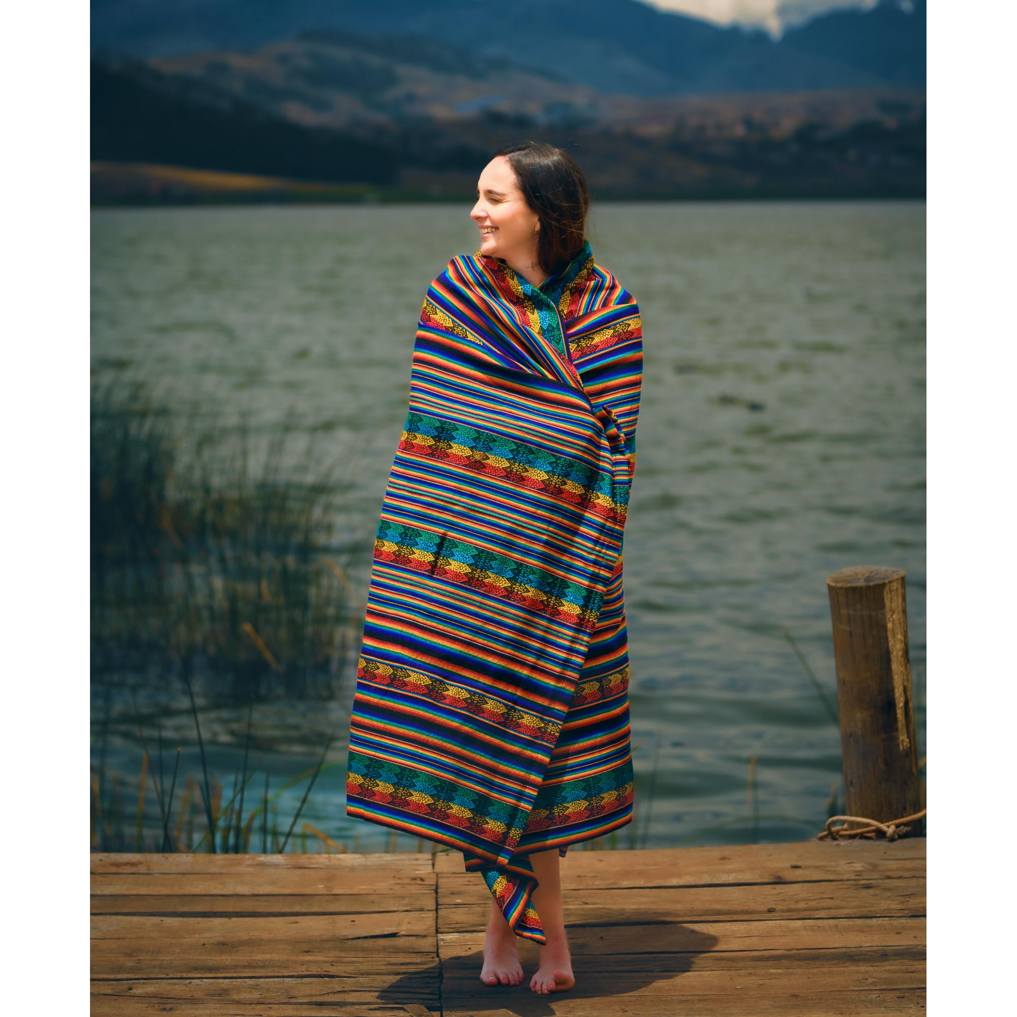 Woman wrapped in a colorful striped blanket standing on a dock by a lake with mountains in the background.