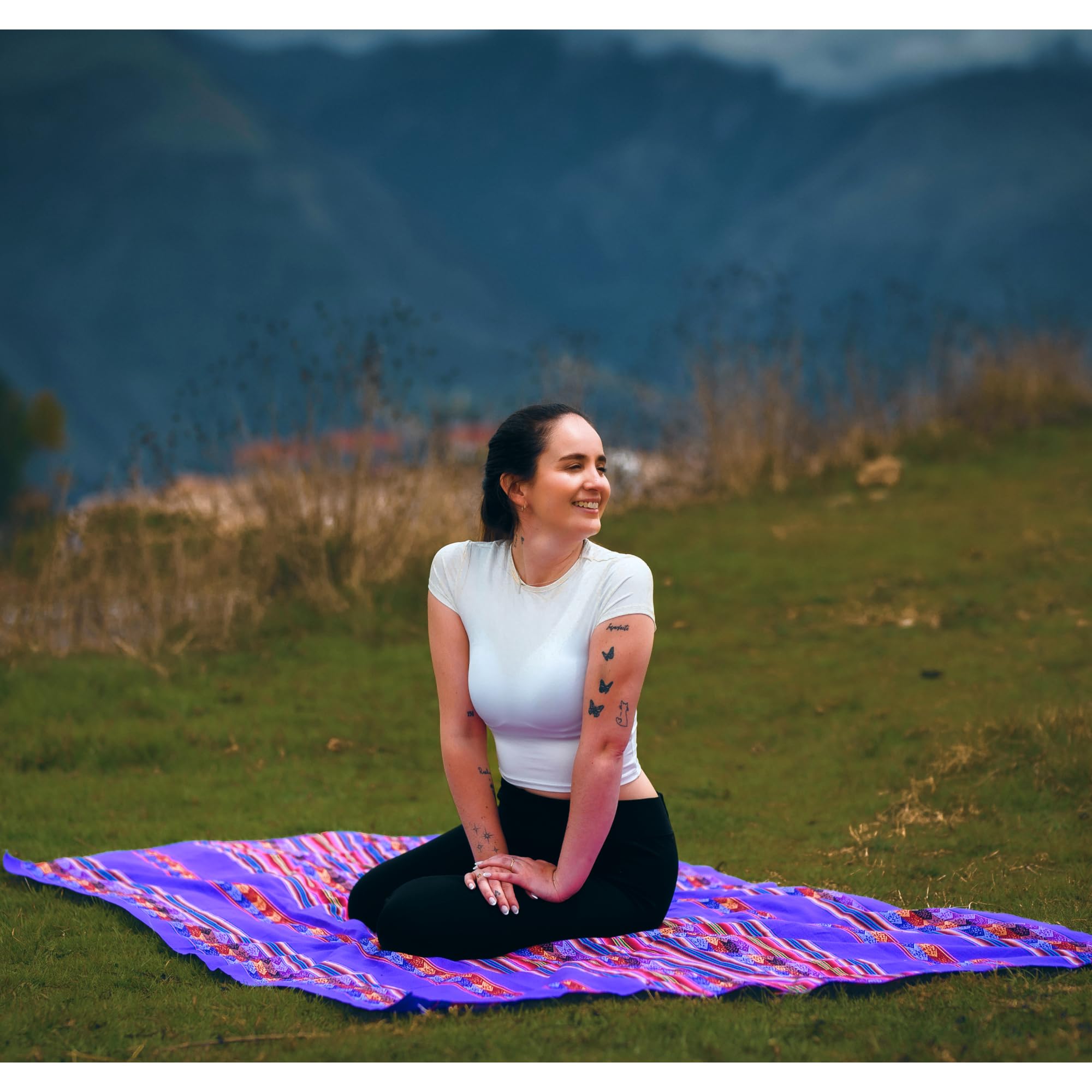 Woman sitting on a colorful blanket in a grassy field with mountains in the background