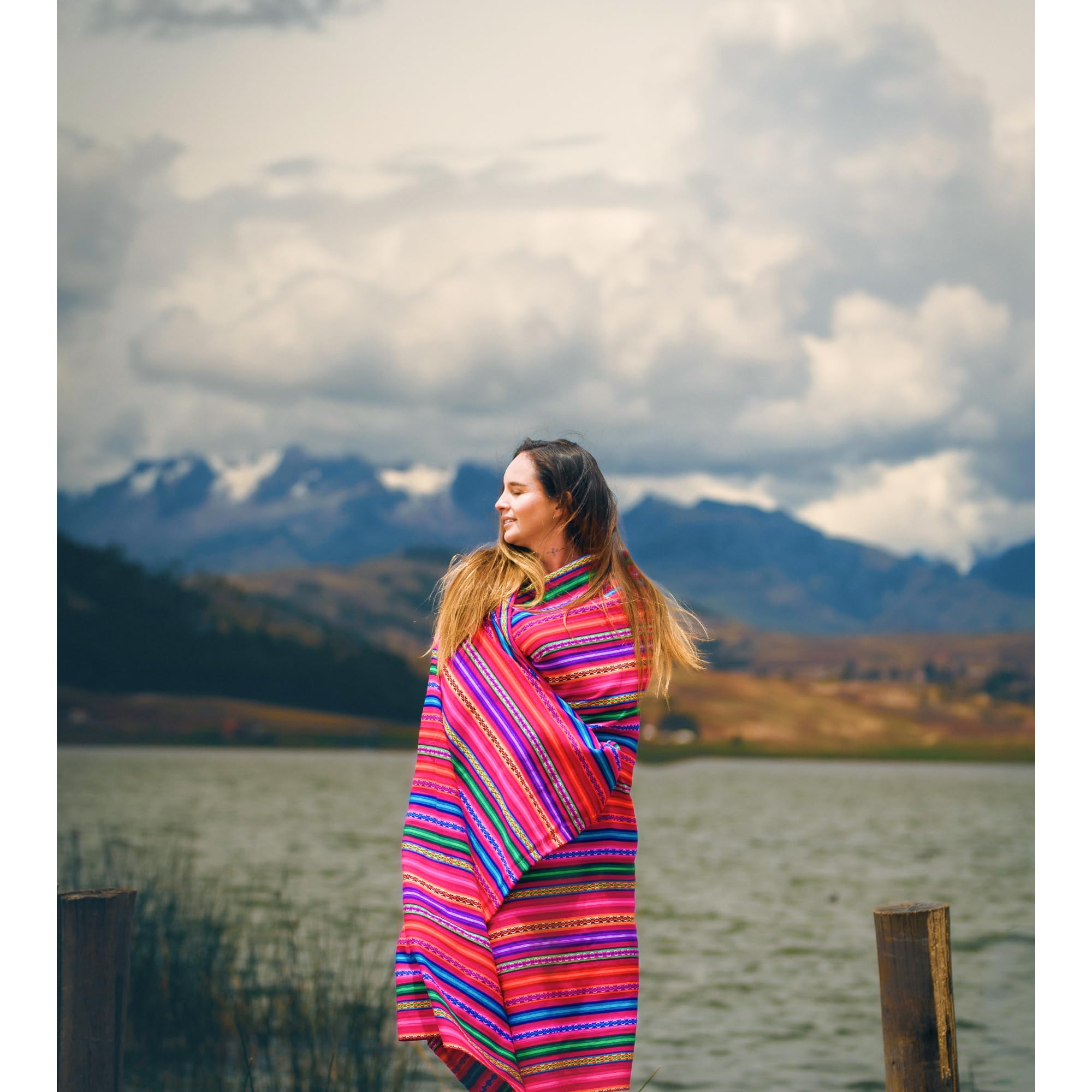 Woman wrapped in a colorful striped blanket standing by a lake with mountains in the background