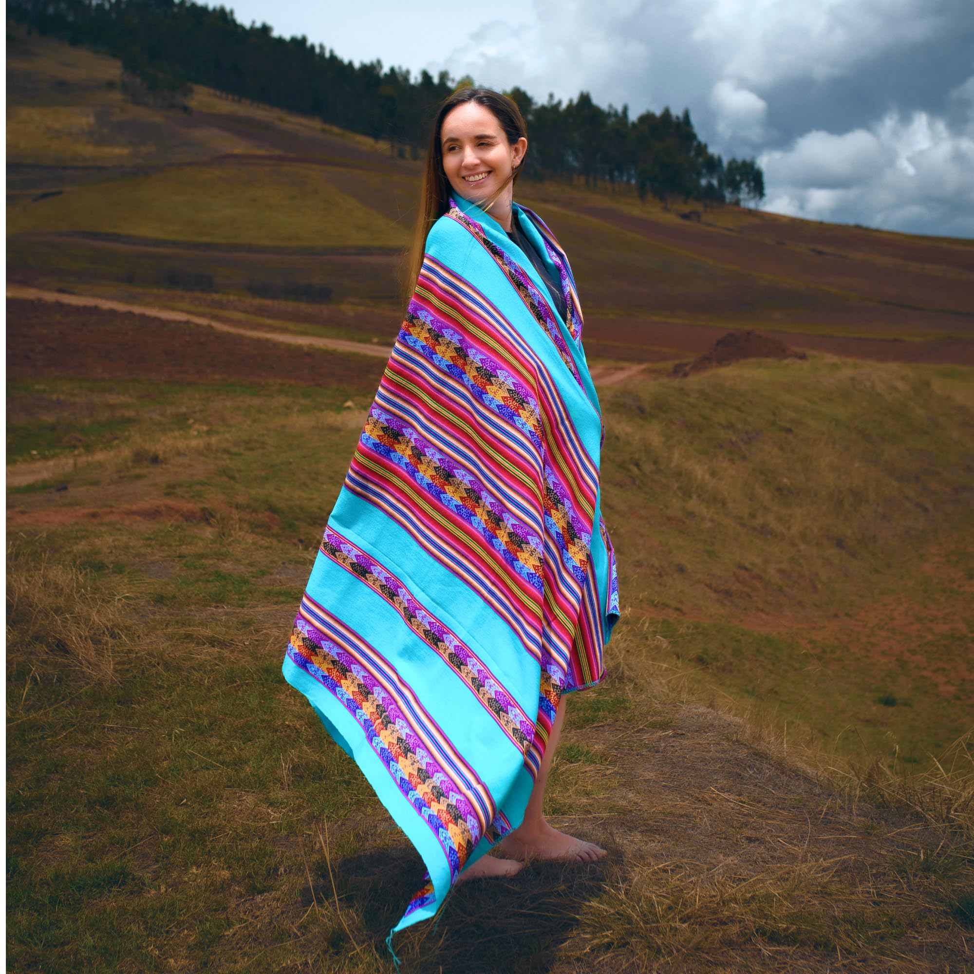 Woman wrapped in a colorful striped blanket standing in a field with hills and trees in the background.