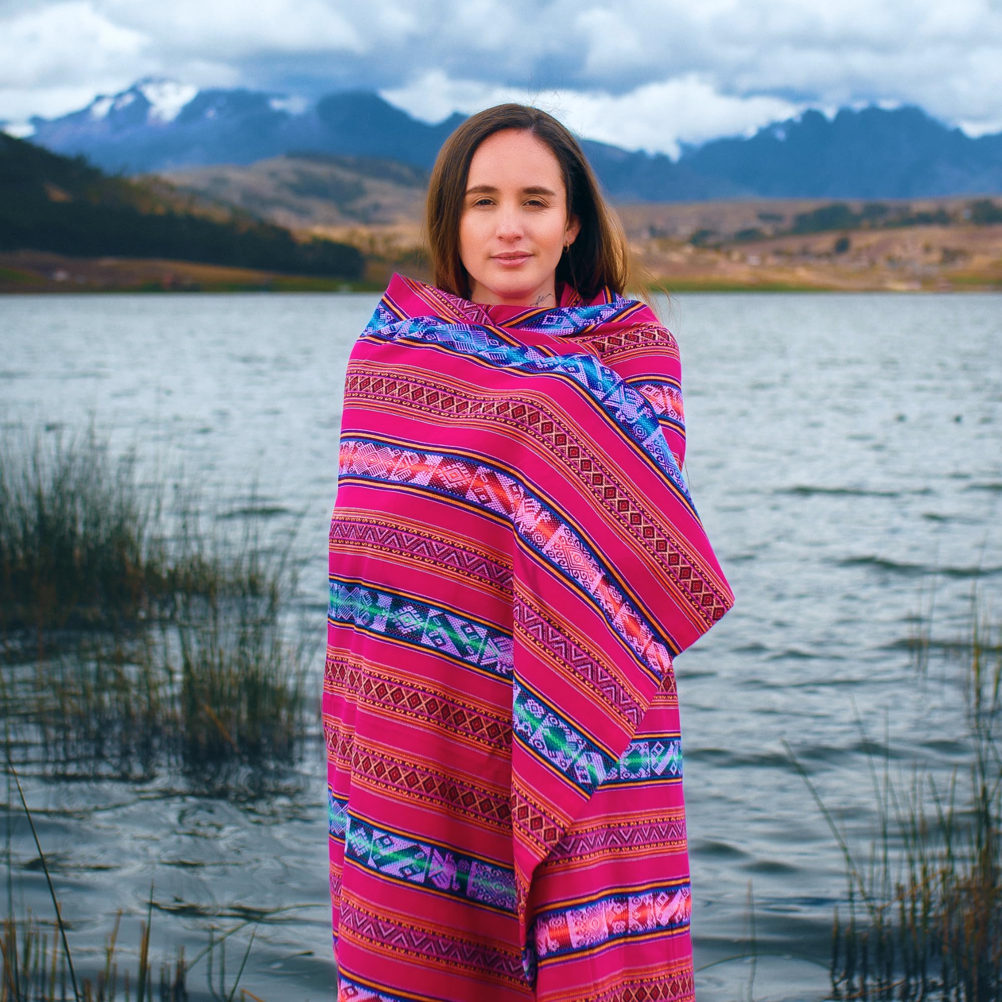 Woman wrapped in a colorful patterned blanket standing by a lake with mountains in the background