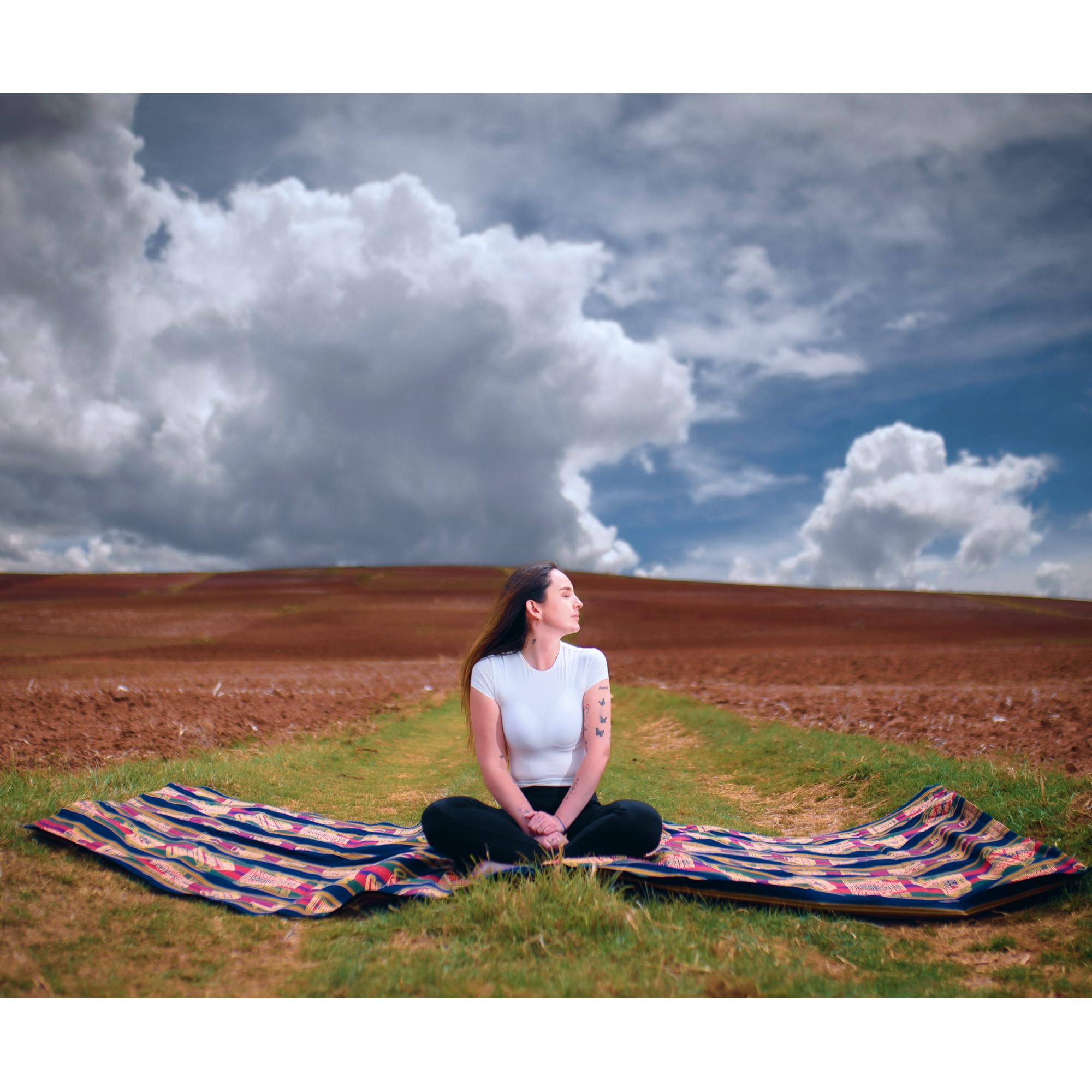 Person sitting on a colorful blanket in a field with a dramatic sky.