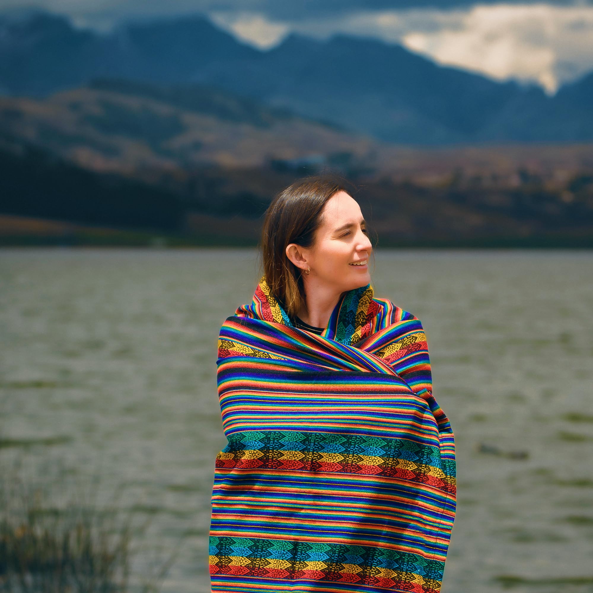 Woman wrapped in a colorful striped blanket standing by a lake with mountains in the background