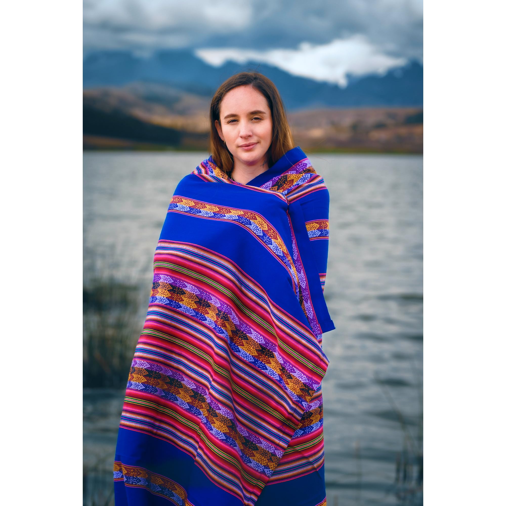 Woman wrapped in a colorful blanket standing by a lake with mountains in the background