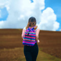 Person wearing a colorful striped backpack in an open field with a blue sky.