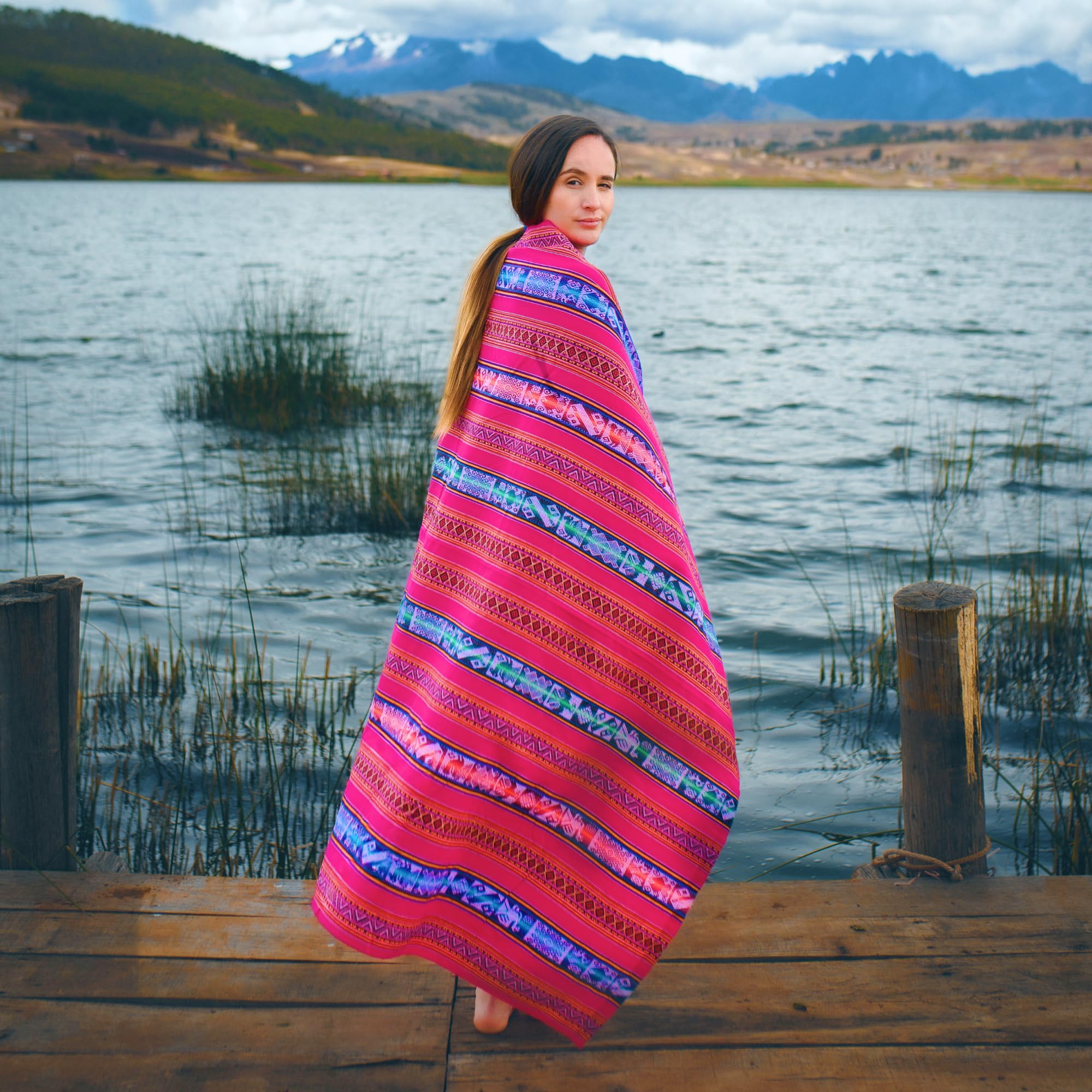 Woman wrapped in a colorful blanket standing on a wooden dock by a lake with mountains in the background