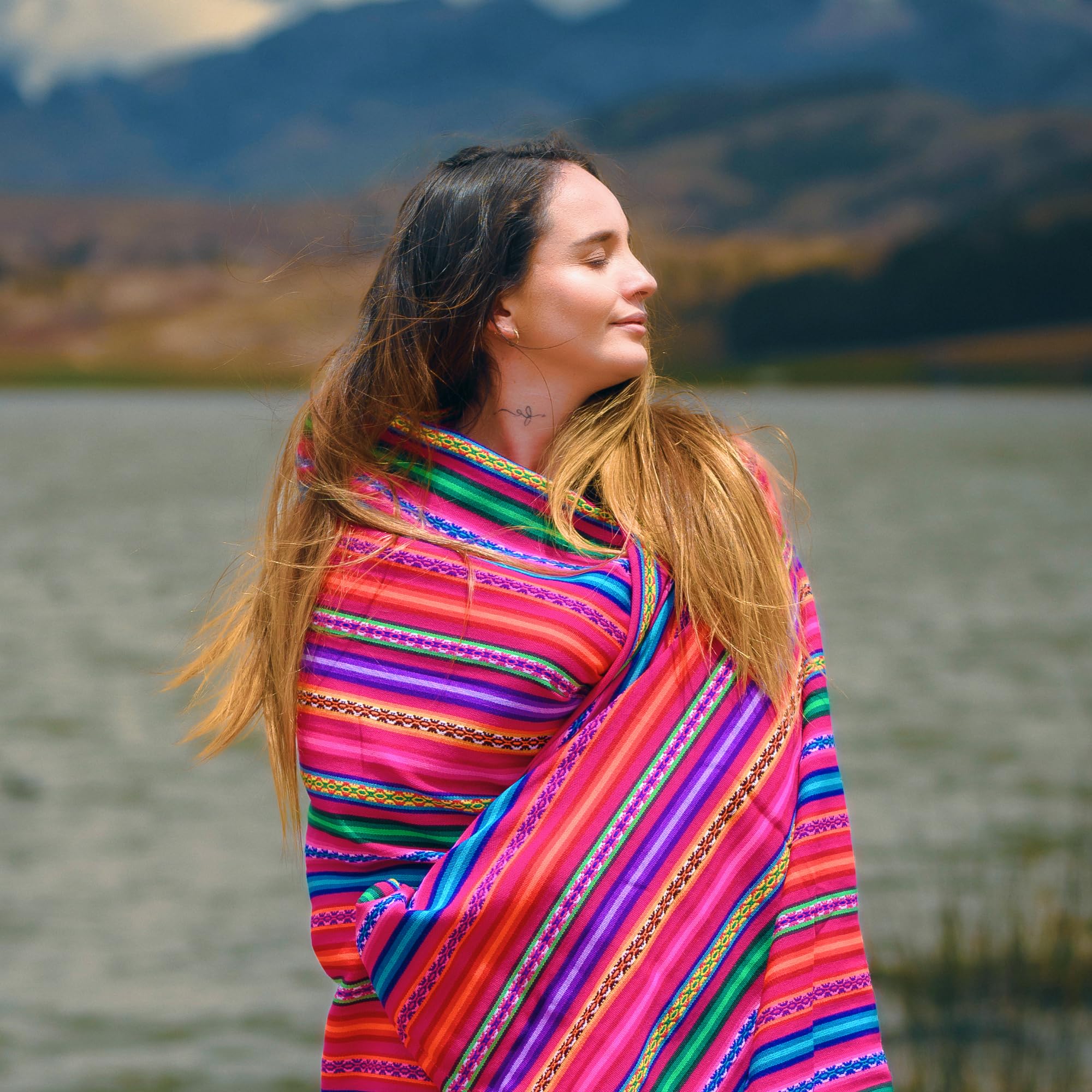 Woman wearing a colorful striped shawl in a natural setting with mountains and water.