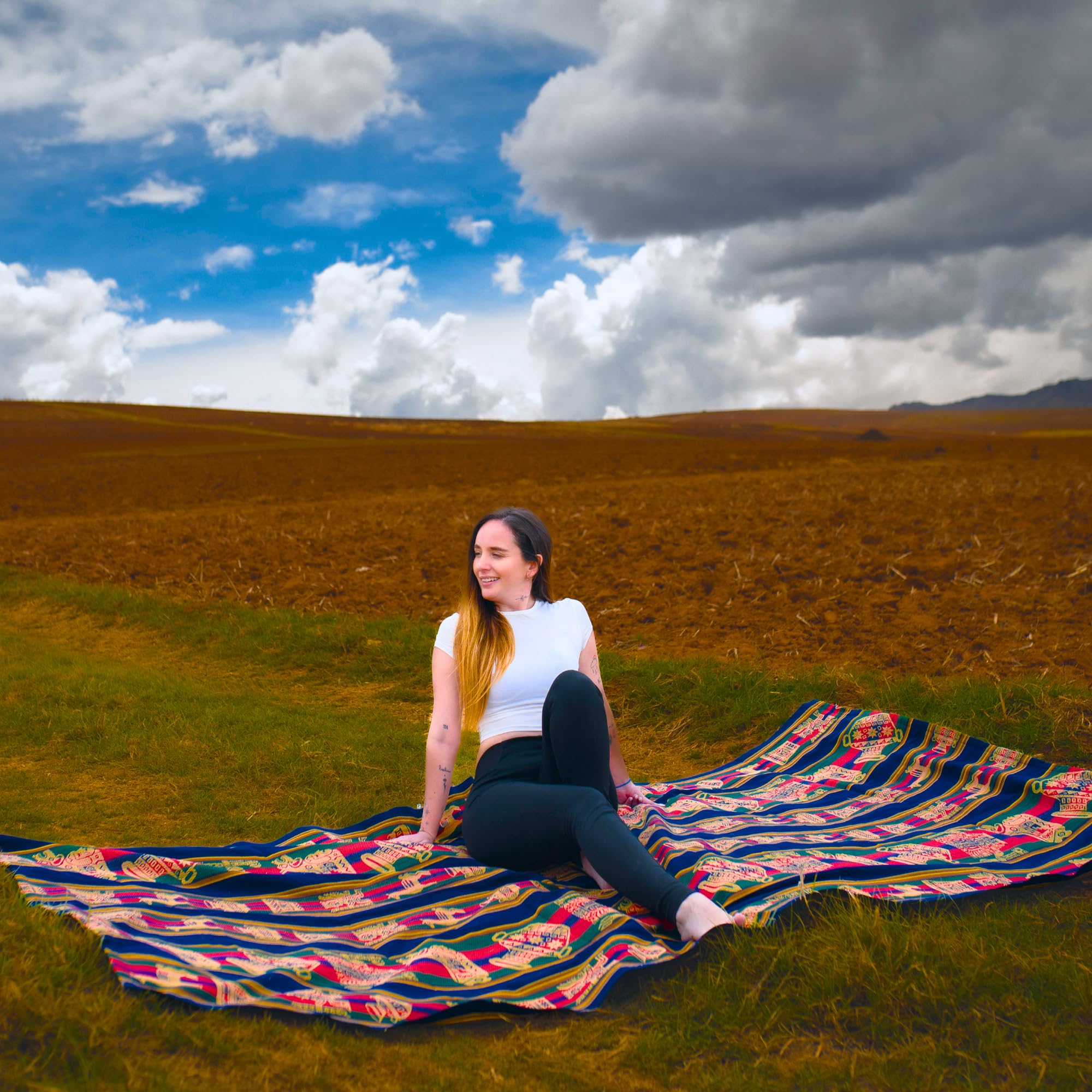 Woman sitting on a colorful blanket in a field with a blue sky and clouds.
