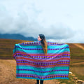 Woman holding a colorful striped blanket in a scenic outdoor setting with mountains and sky.